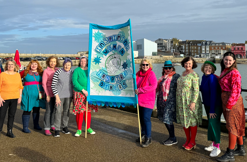 An image of WI members on the harbour arm holding a banner, they are in bright spring colours.